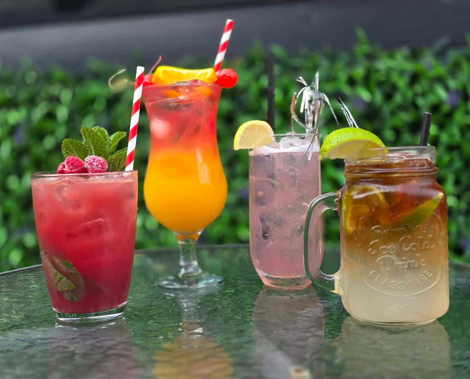Four cocktails in tall and mason-jar glasses on a glass table by a green hedge, including a raspberry mojito